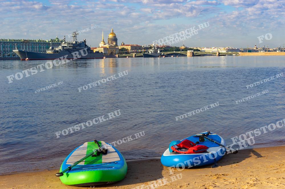 На фотографии изображён летний день на берегу Невы в Санкт-Петербурге. На переднем плане — доски для сапбординга на песчаном берегу. Вдали виден Эрмитаж и Исаакиевский собор, а также пришвартованный военный корабль. Мост и архитектурные памятники подчёркивают величие и красоту города.